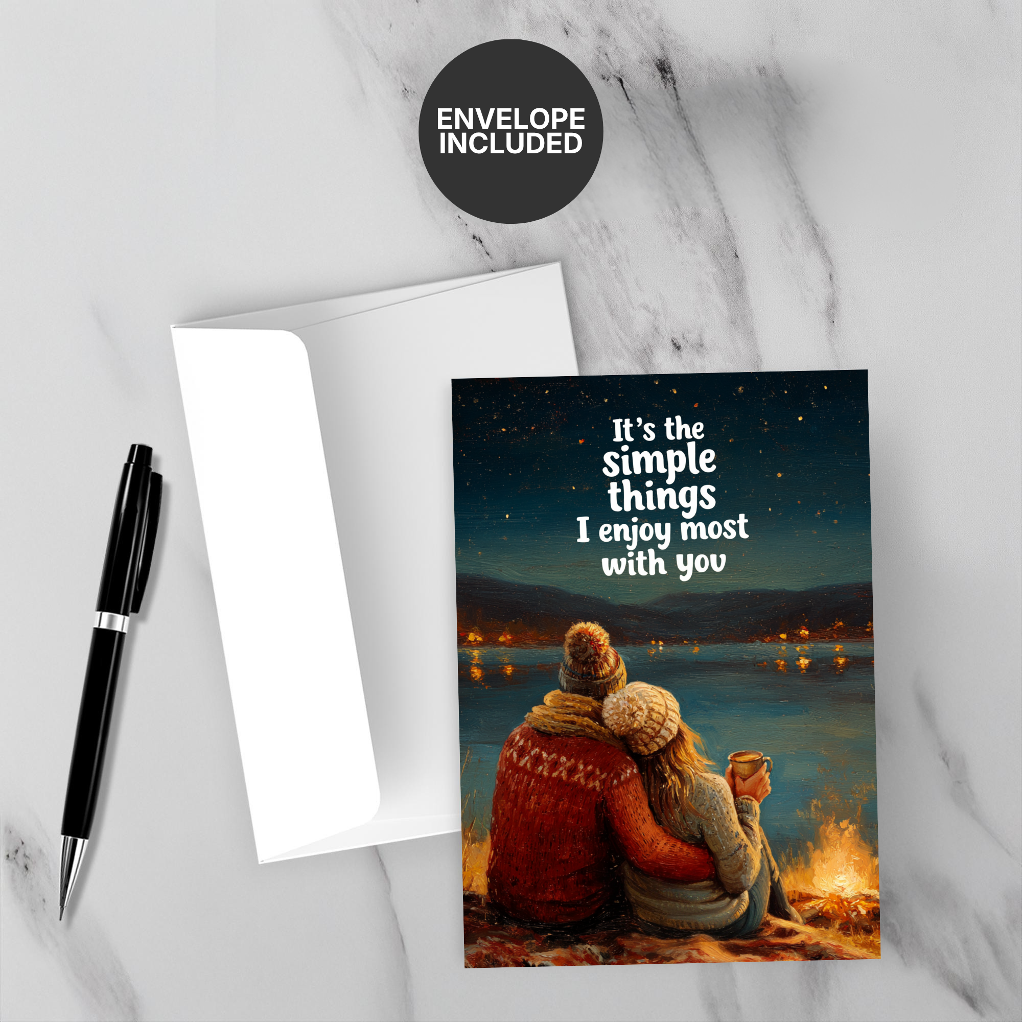 Greeting card with a couple by a campfire and text 'It's the simple things I enjoy most with you' on a marble surface with a pen and envelope.