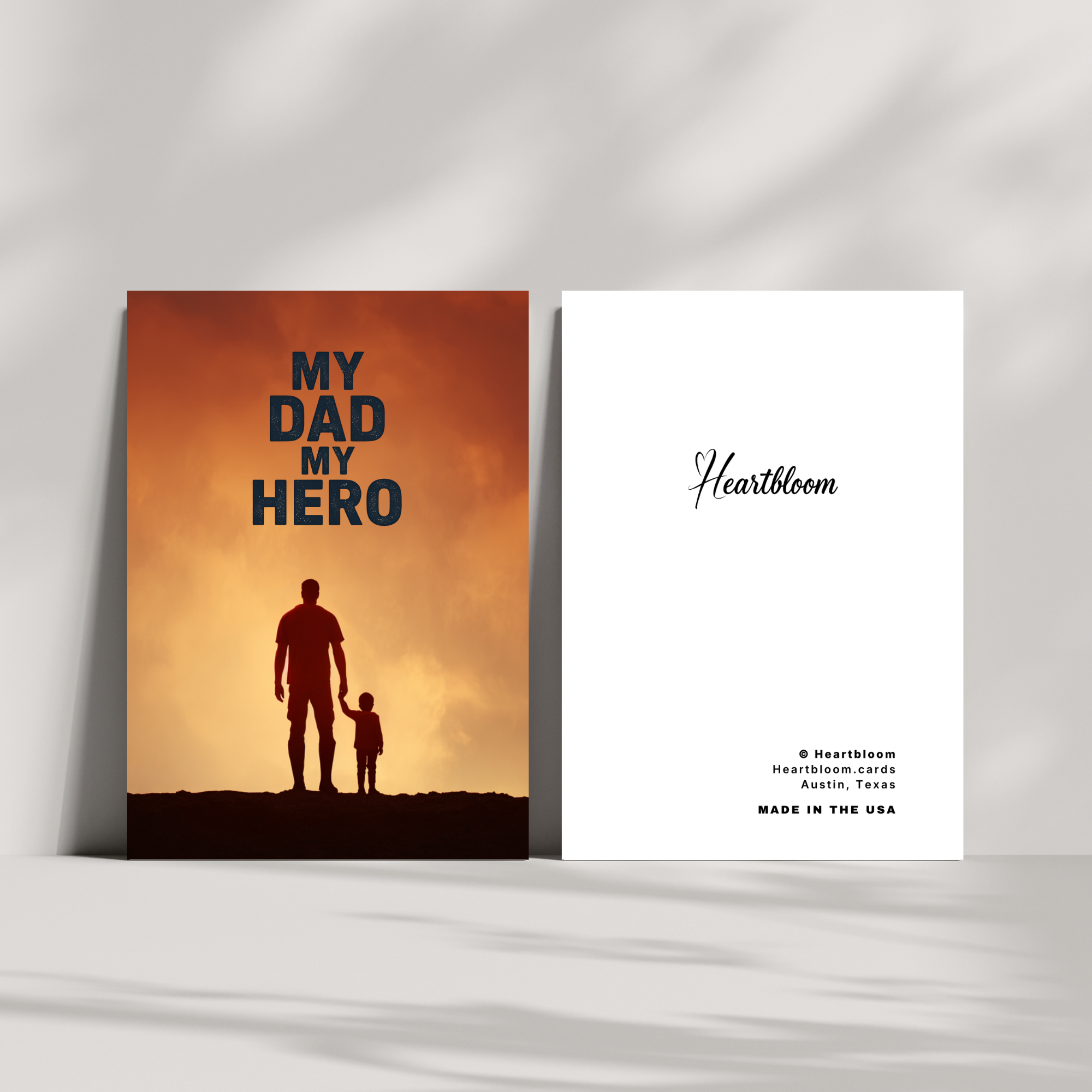 Card with silhouette of a father and child against an orange sky, featuring the text 'My Dad My Hero' and 'Heartbloom' branding.
