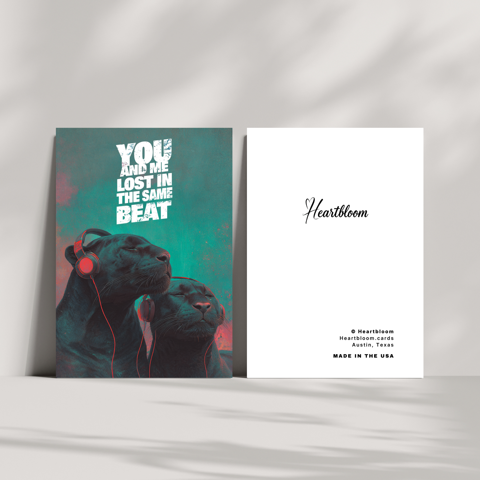 Two greeting cards on a white surface, two black panthers wearing headphones and the other with 'Heartbloom' branding.