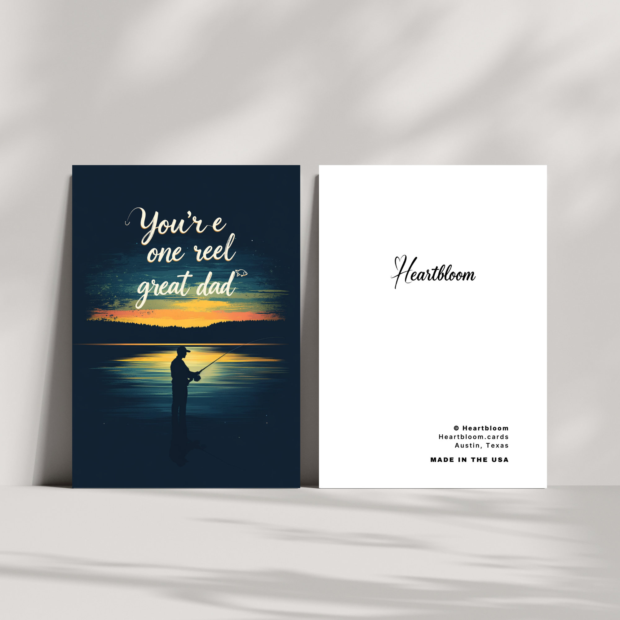 Two greeting cards on a white surface, one with a sunset fishing scene and the other with 'Heartbloom' branding.